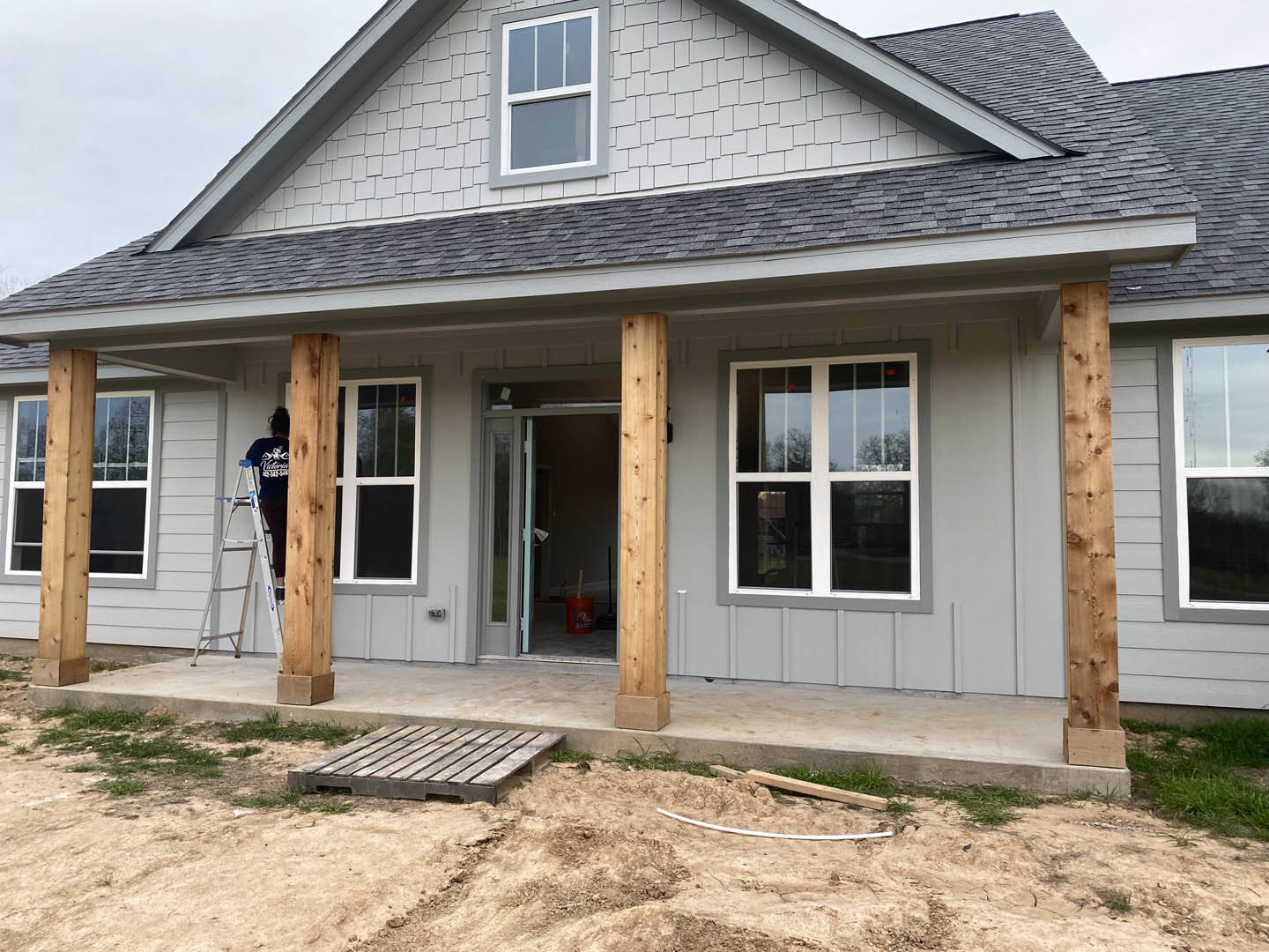 Person on a ladder installing white-framed window on house exterior with gray siding, wooden pallet on ground, covered porch visible