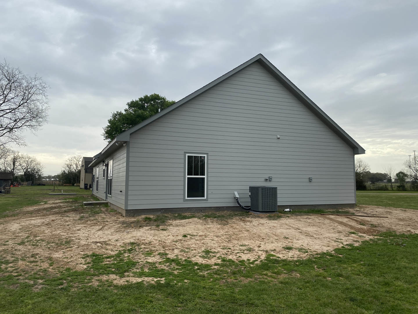 Grey house with white-trimmed window, fenced grassy yard, leafless tree in background, cloudy sky overhead
