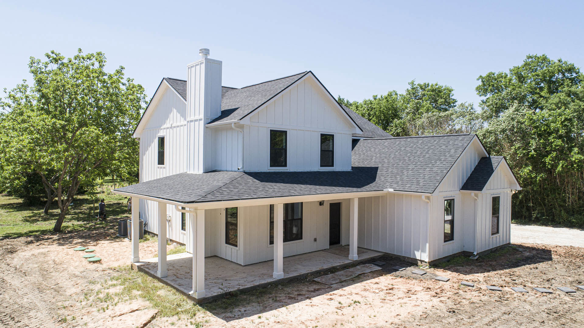 White house with black roof, black and white window frames, white pillars on porch, dirt path alongside, tree and people in background