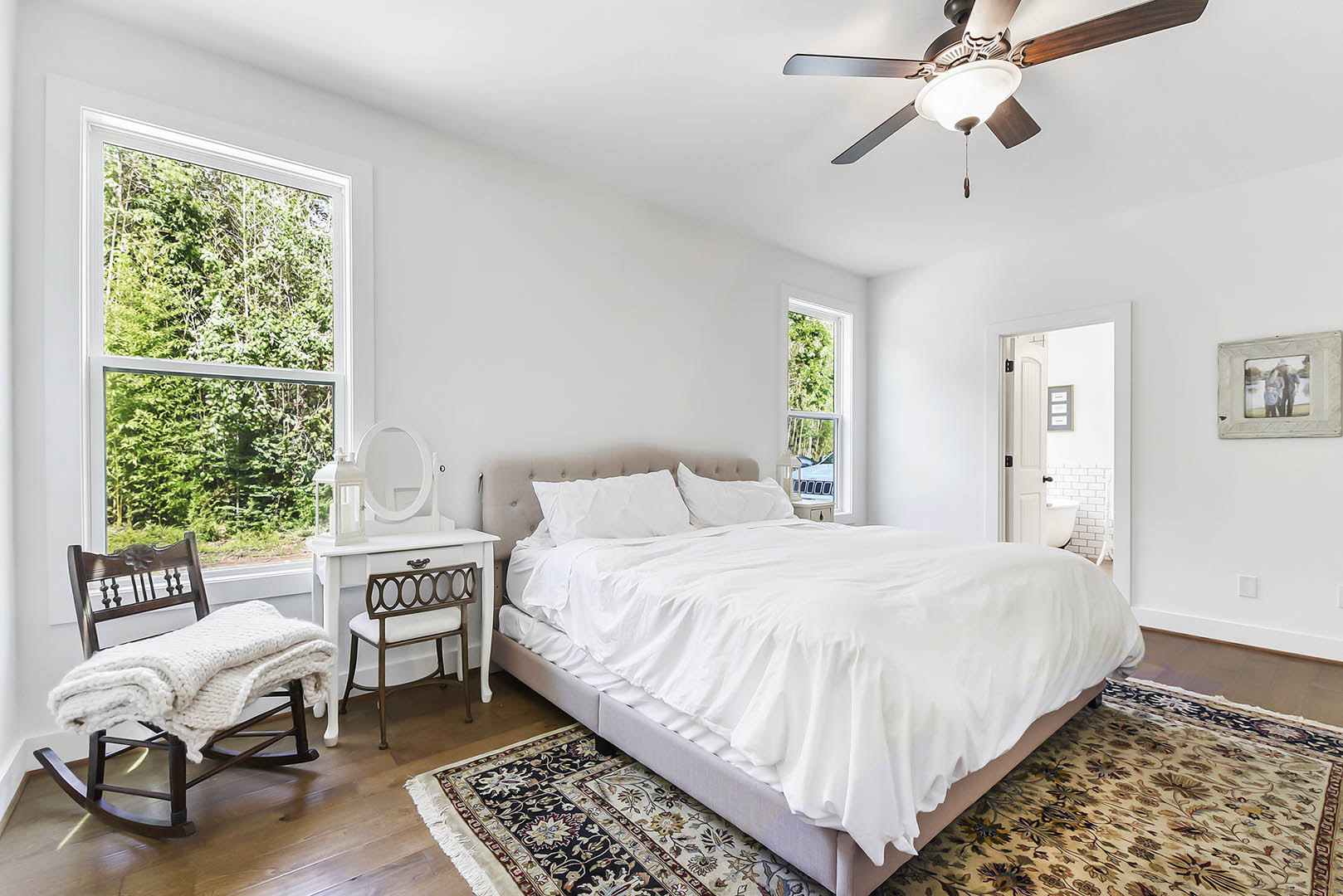 Bedroom with a bed dressed in white sheets and pillows, ceiling fan with light fixture, rocking chair draped with a blanket, picture frame showing two people, and chair with white