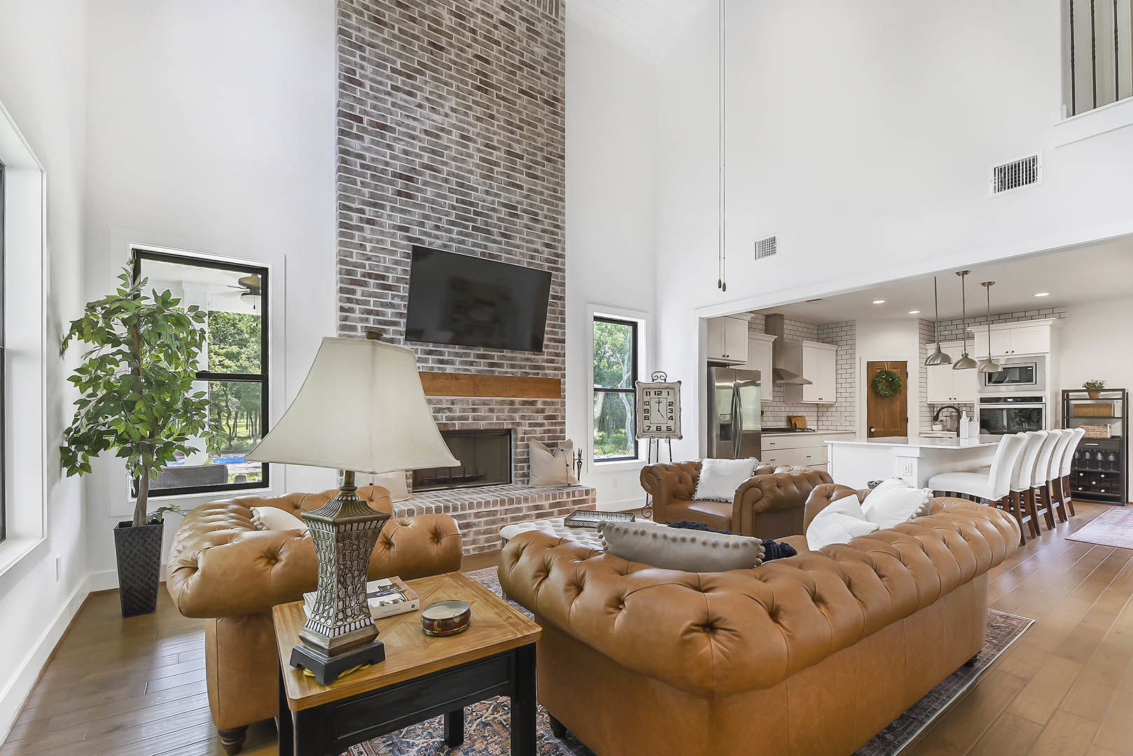Living room with exposed brick fireplace, black screen above mantel, brown leather couches with pillows, potted tree, round metal container, and white lamp shade.