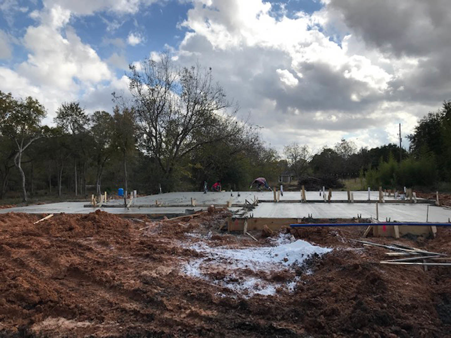 Dirt construction site bordered by tall trees under a cloudy sky, patches of snow scattered on the ground