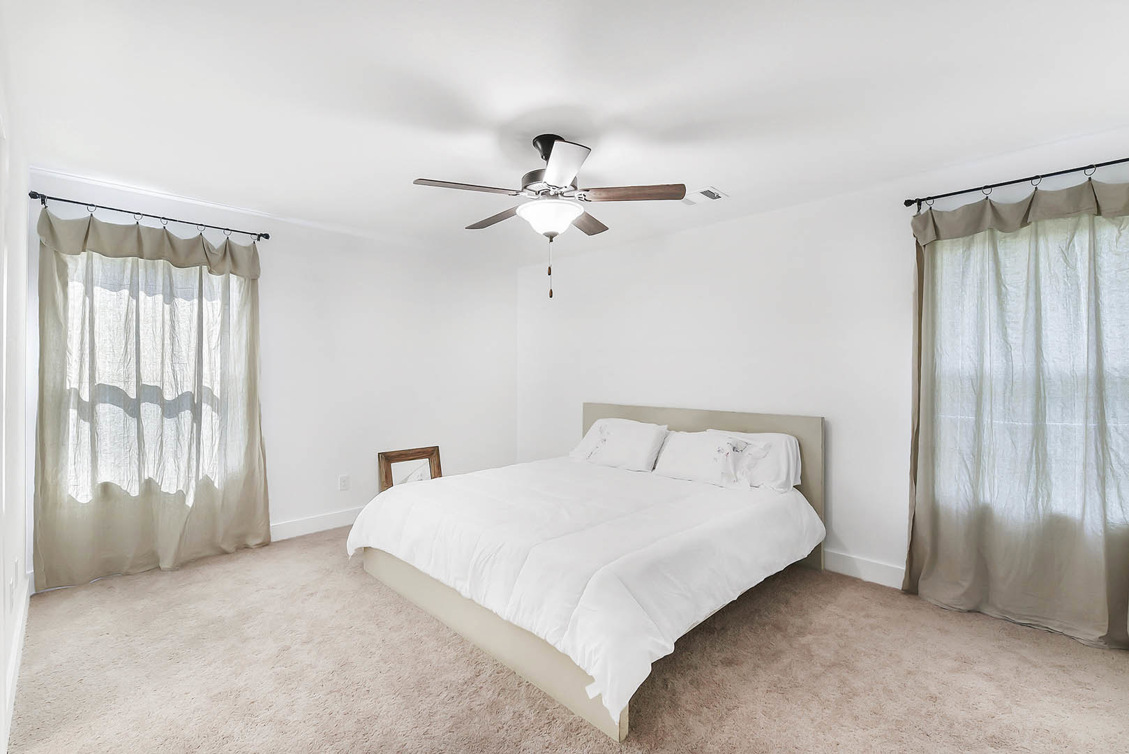 Queen bed with white sheets and pillows, white curtain filtering light on window, ceiling fan with light fixture above, neutral walls and minimal decor