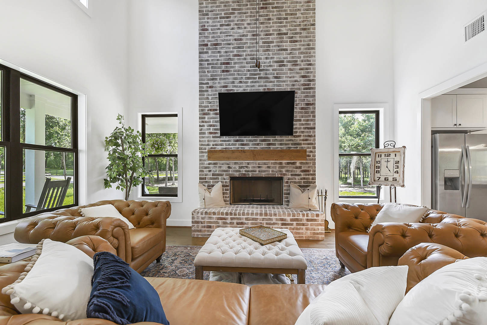 Living room with a brick fireplace, mounted black TV, brown leather couch with white pillows, white ottoman holding a tray, and large window in the background