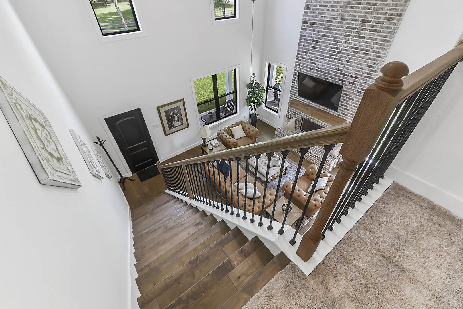 Open living area with wood staircase, gray couch facing stone fireplace, framed artwork on wall, black door, brown leather chair with pillow, outdoor table and chair visible