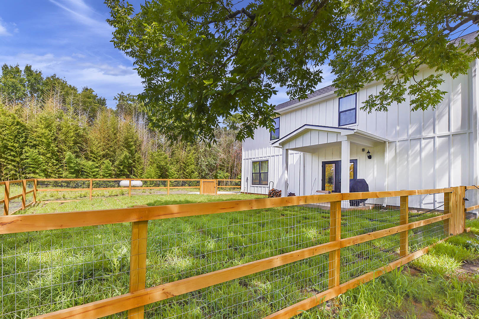 White split rail fence surrounding a grassy yard, wooden gate open near porch, white house exterior with large windows, leafy tree branches overhead, blue sky with scattered clouds