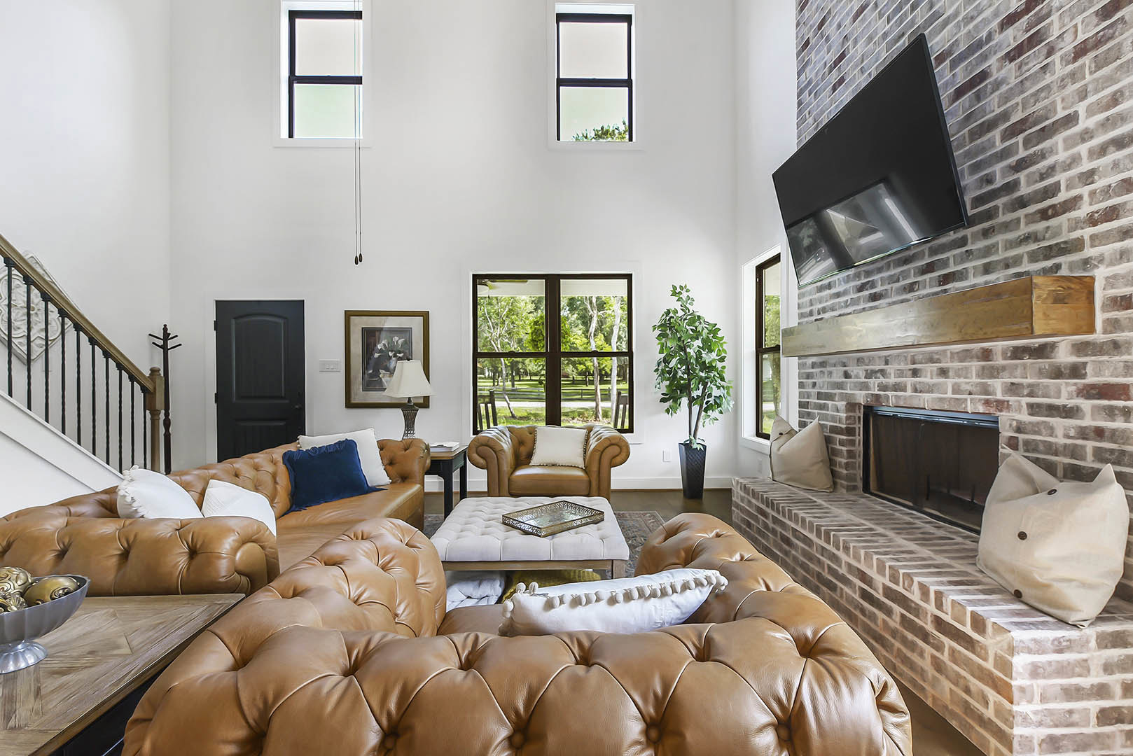 Living room with brown couch, white pillow on leather chair, brick fireplace wall, black television, silver tray on coffee table, and green potted plant