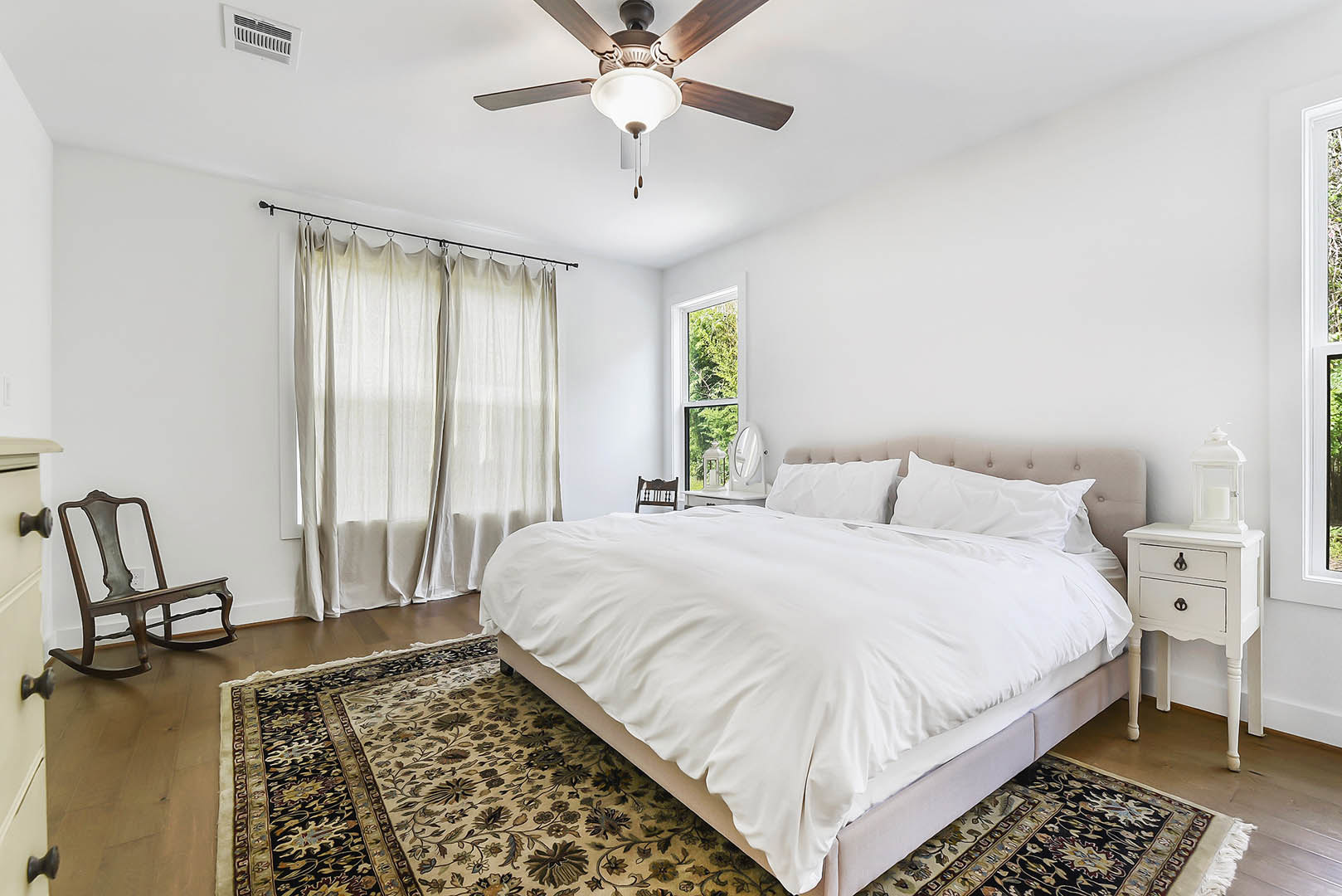 Bedroom with a large bed featuring white sheets and pillows, patterned rug, wooden rocking chair, white nightstand with a clock, white curtains, and ceiling fan with light fixture