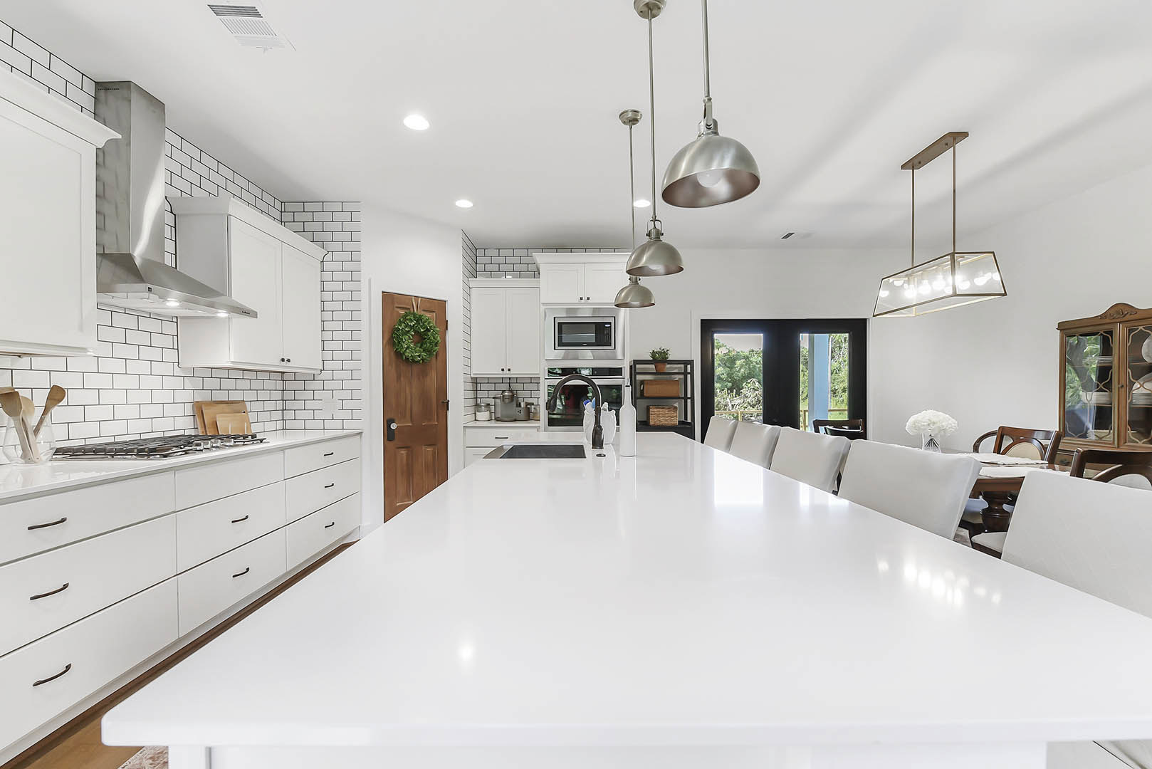 White kitchen featuring white cabinetry and countertops, central island with black faucet, modern ceiling light fixture, adjacent room with white couch and sofa, green wreath