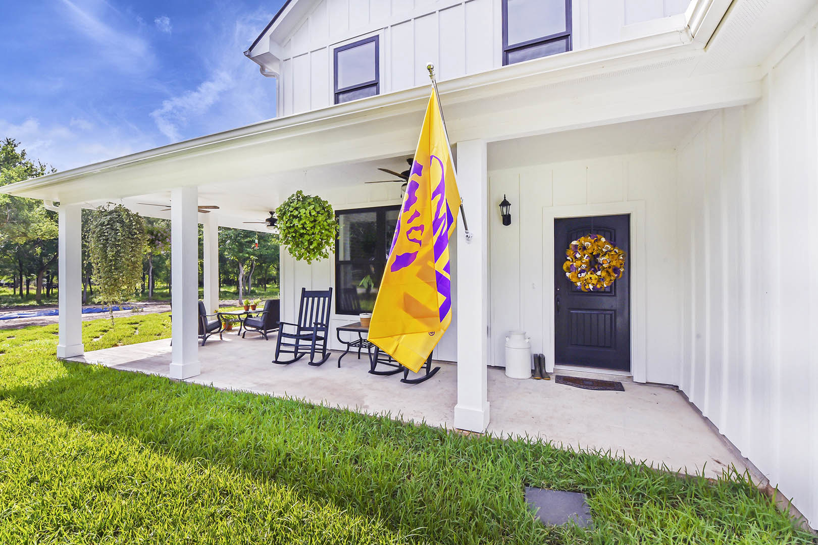 White siding house with a yellow and purple flag hanging from the front porch, wreath on the door, white rocking chair, white handled container, and potted green plant.