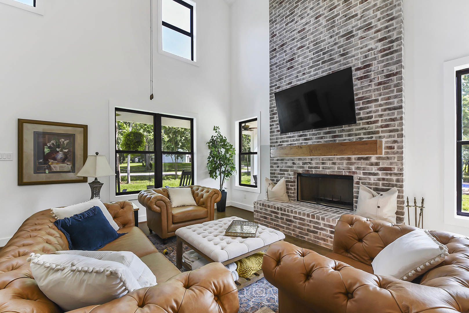 Living room with brown leather sofa and chair, brick fireplace, black television mounted on brick wall, white pillow, still life painting, potted plant, patterned metal tray, white