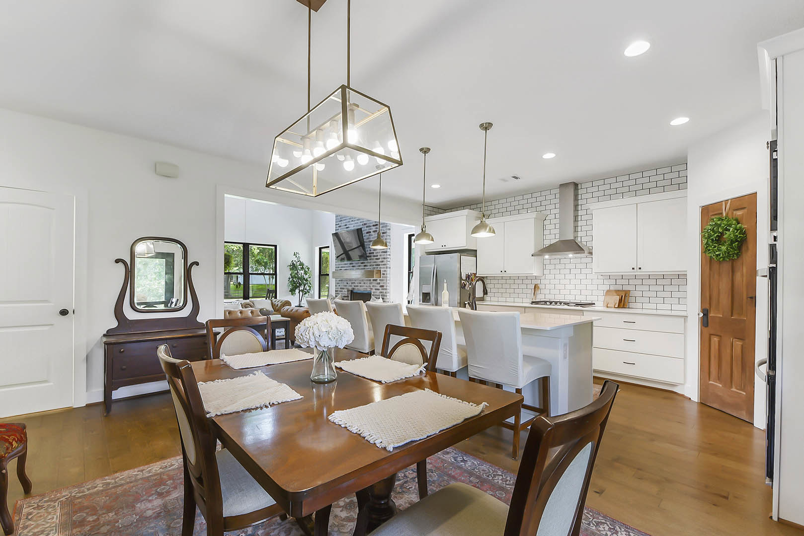 Open dining room and kitchen featuring a large wooden table with white placemats, glass vase of white flowers, modern glass box light fixture, neutral cabinetry, and hardwood