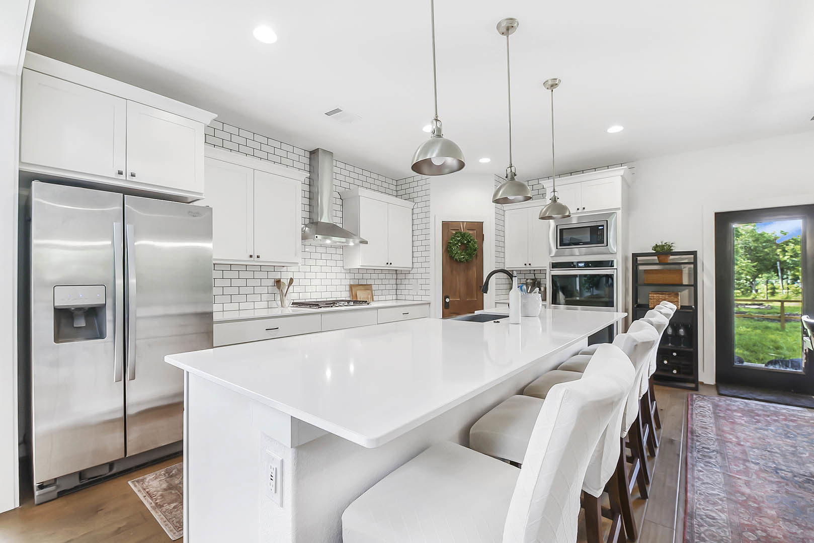 White kitchen with shaker cabinets, quartz countertops, stainless refrigerator, island with bar stools, window overlooking fenced yard, green wreath on door, light carpet flooring