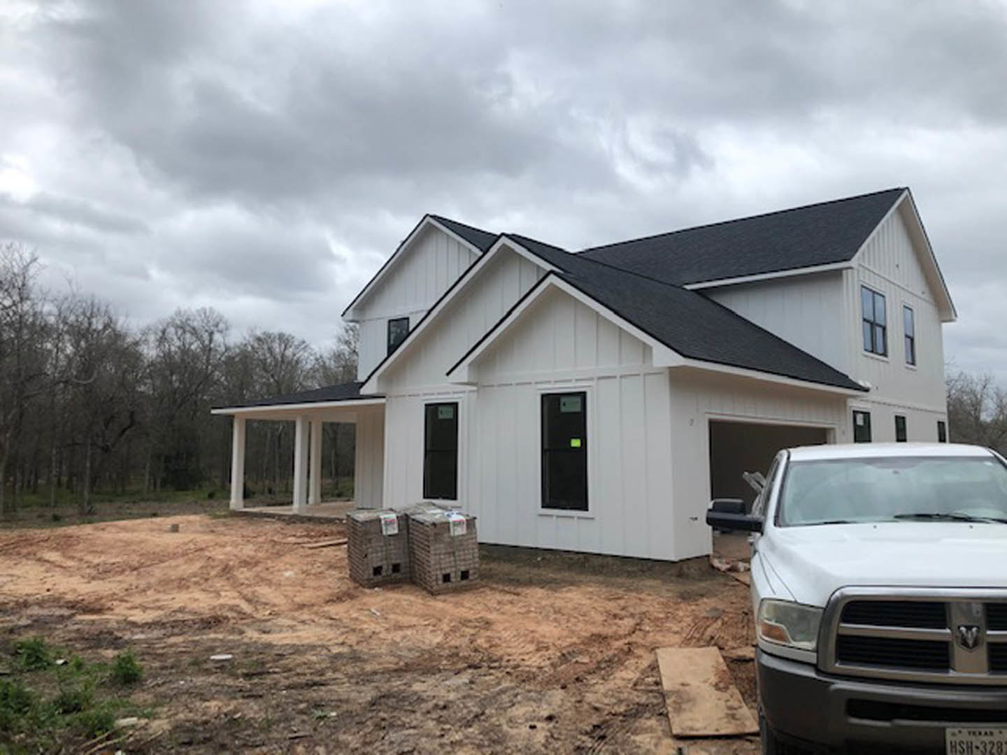 White house with black roof under construction, white truck parked on dirt, pallets and labeled boxes stacked near foundation, cloudy sky overhead.