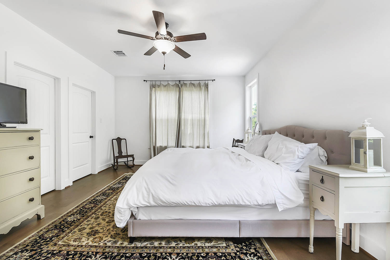 Bedroom with white sheets and pillows on the bed, ceiling fan with light fixture, curtain, corner chair, and chest of drawers