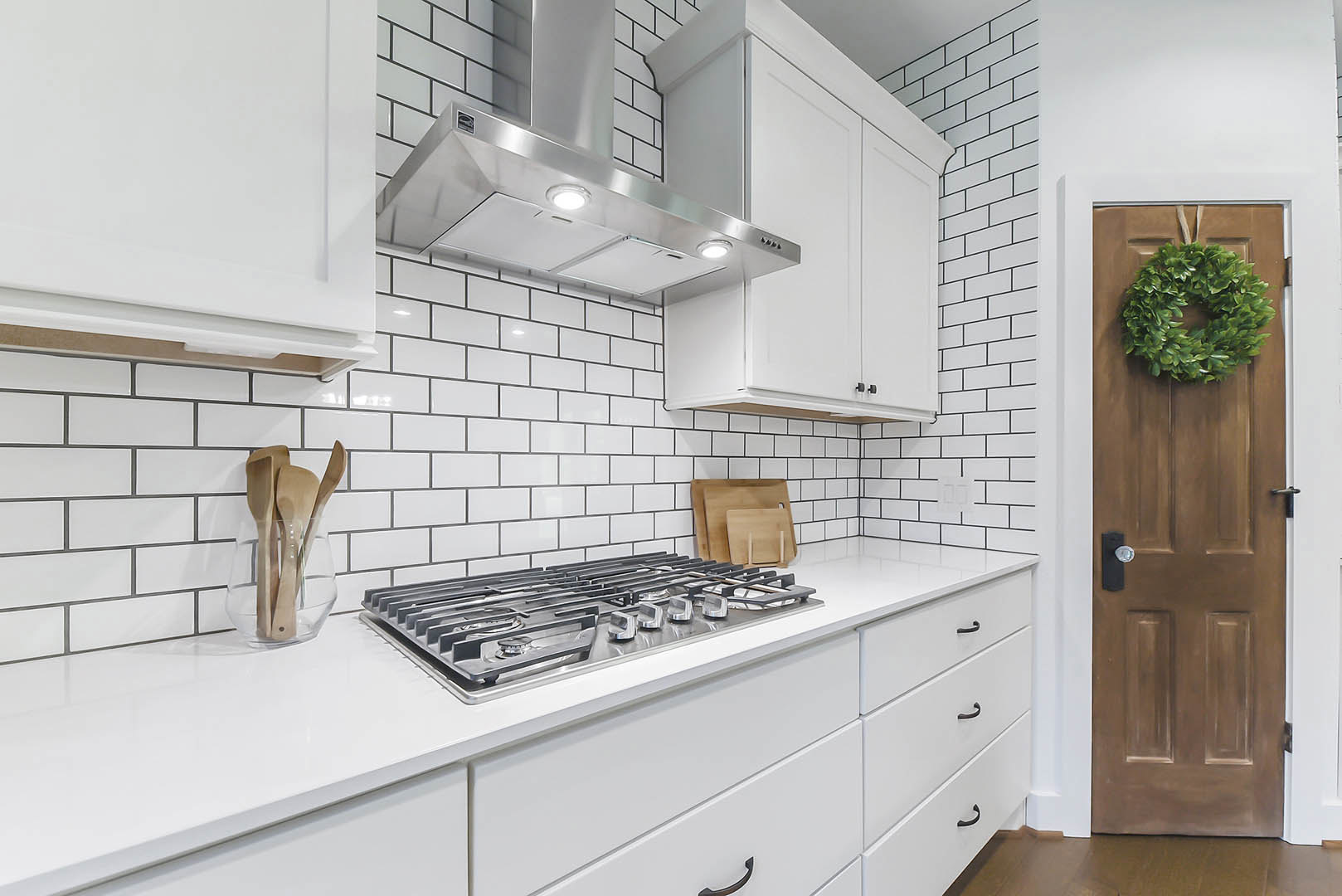 White kitchen with shaker cabinets, quartz countertops, stainless steel sink, wooden spoons in a glass vase, green wreath hanging on a door, close-up views of stove top and vent
