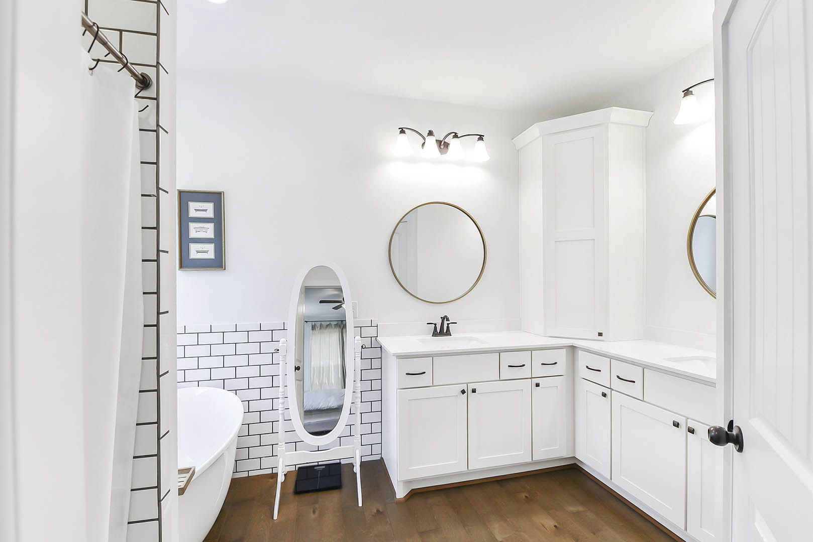 Bathroom with a circular wall mirror above a white vanity, freestanding white bathtub, framed pictures on light-colored walls, and a close-up of a modern light fixture.