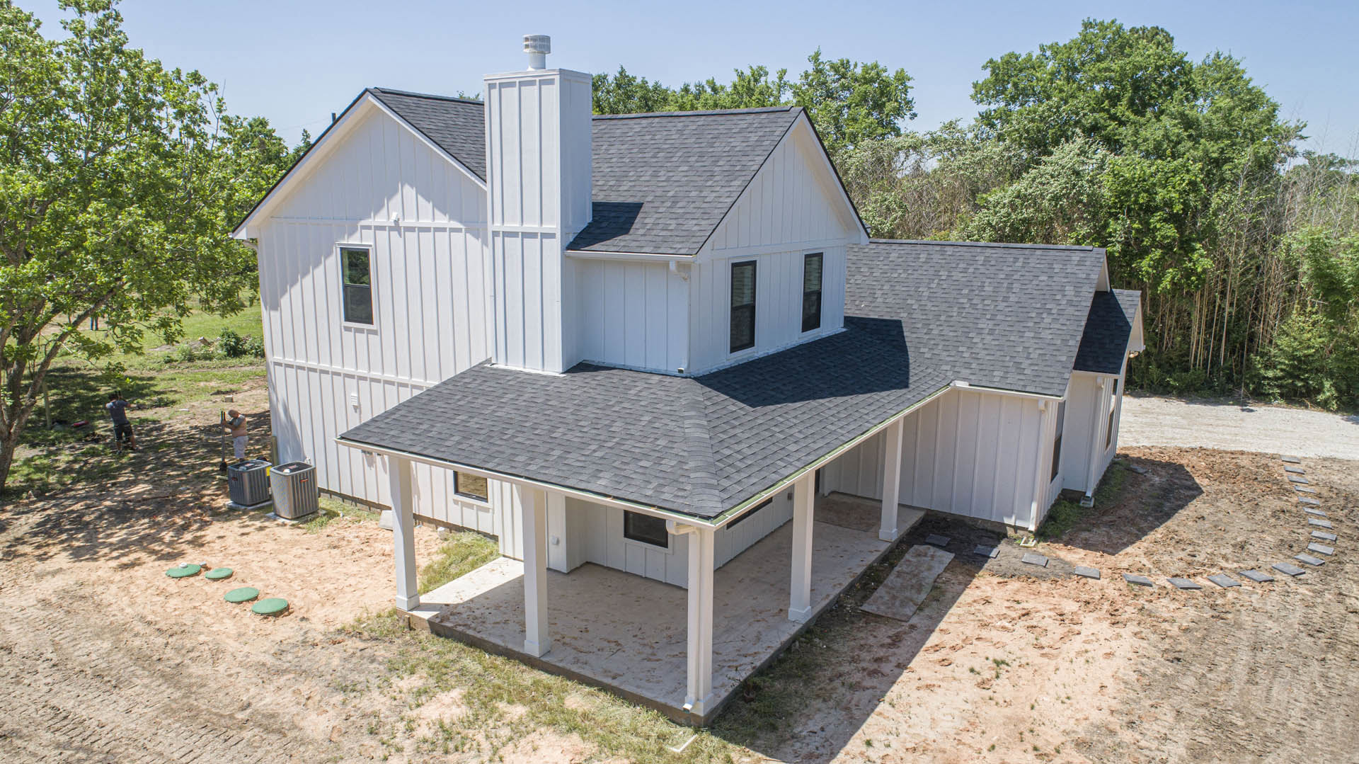 Wide front porch with white columns, dark wood decking, and large windows framed in white; surrounded by mature trees and landscaped yard under a clear sky