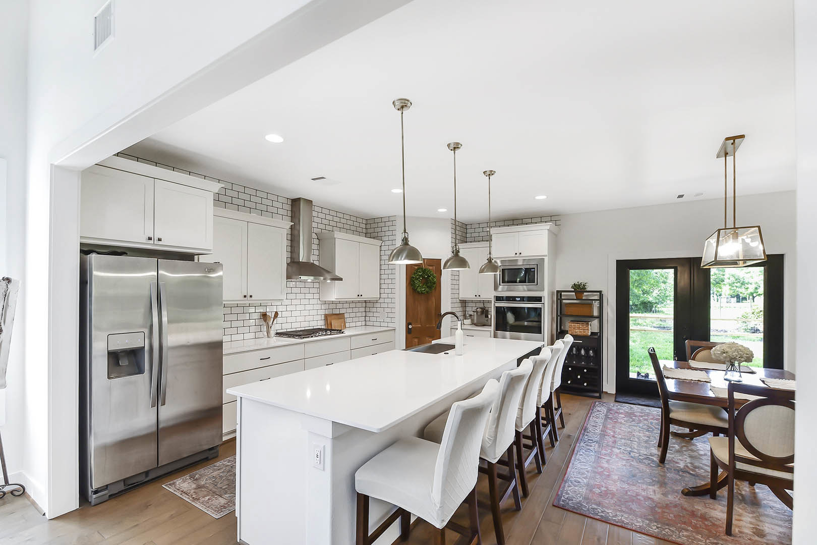 Spacious white kitchen featuring a large central island with white countertops and chairs, oversized stainless steel refrigerator, pendant light fixture, green wreath on door, and