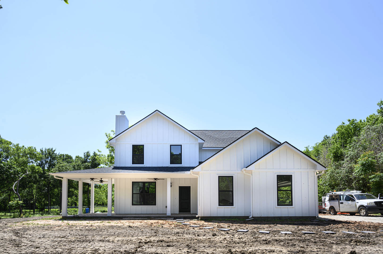 White house with black-framed windows and black front door, surrounded by dirt construction area, white truck with ladder parked nearby, blue sky visible through window, tree in