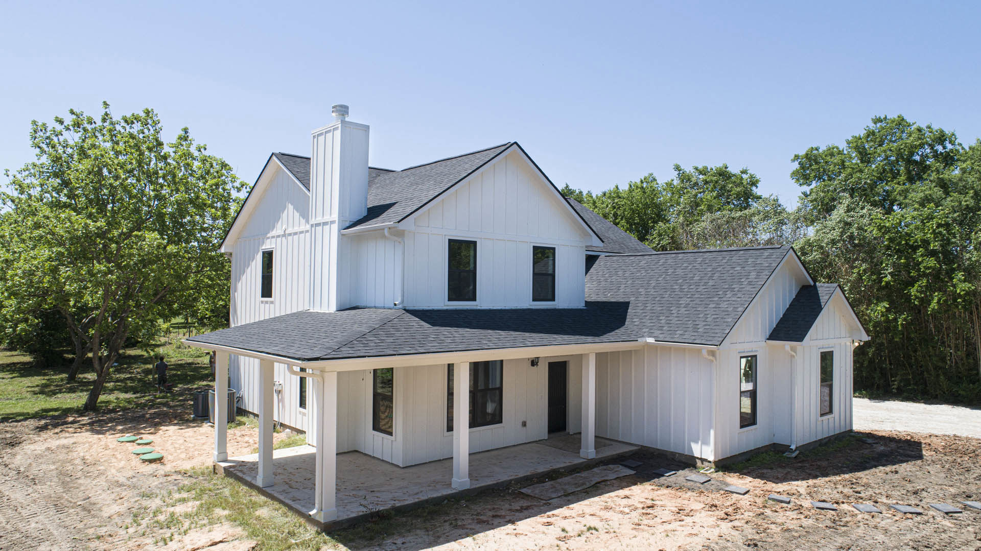 White siding house with black shingle roof, white-framed windows, black front door with white trim, green-leafed tree in front yard, concrete sidewalk, blue sky overhead