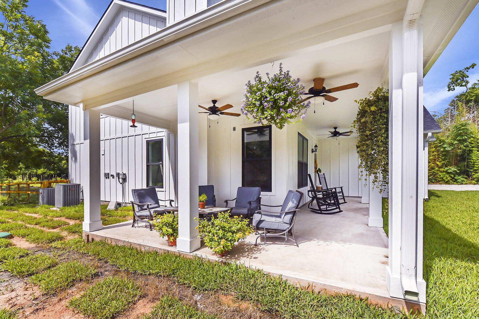 White siding home with black-framed windows, front lawn featuring yellow flowering plants, patio area with chairs and table under shaded porch.