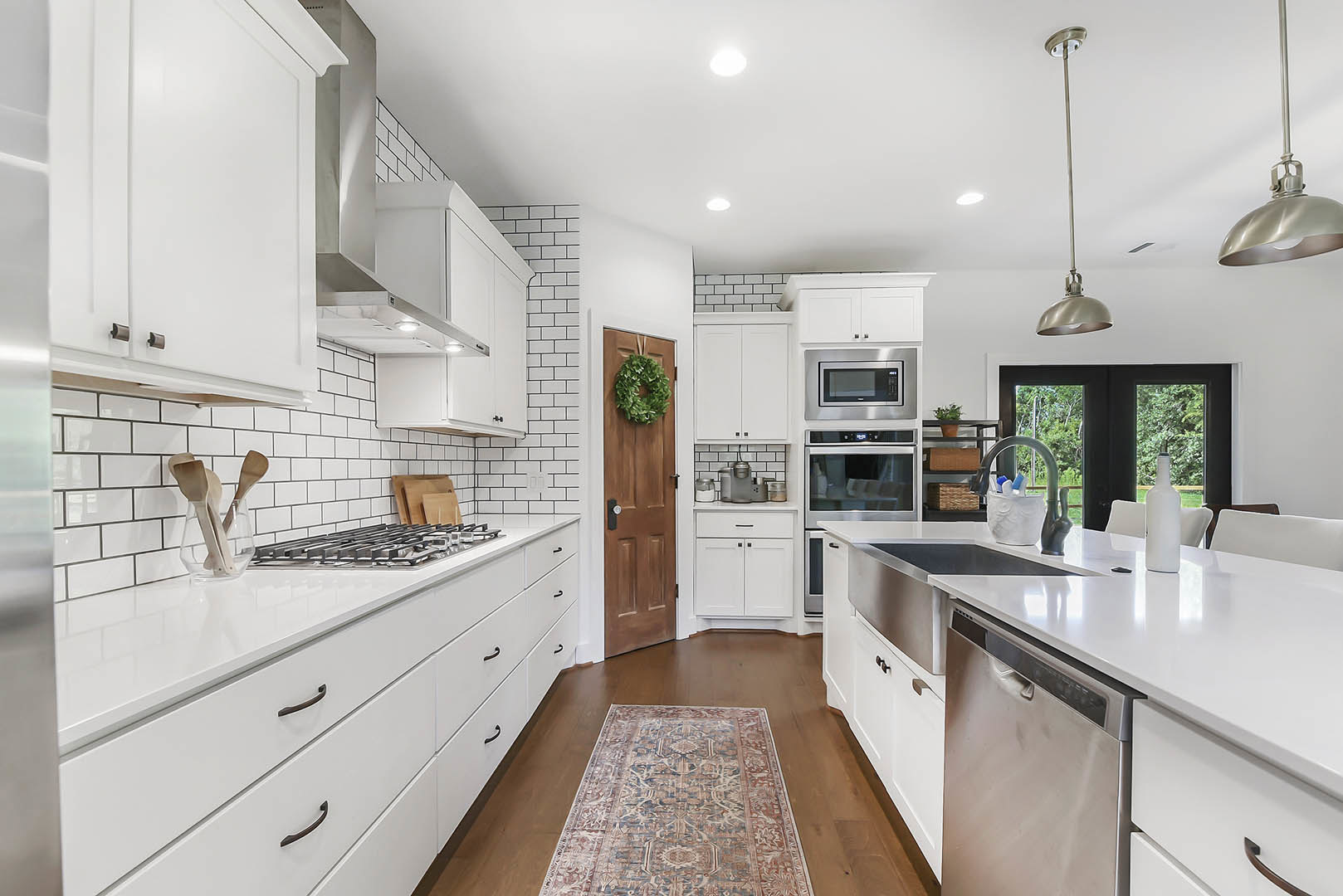 White kitchen cabinets, patterned rug on tile floor, wooden spoons in glass holder on countertop, black microwave oven, tall metal light fixture, stainless steel sink, neutral