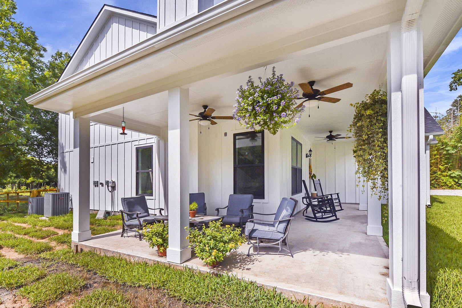 White siding house with covered porch, two wooden chairs, potted purple and yellow flowers, cup resting on chair, lit window, front door, shaded entry, green trees in background.