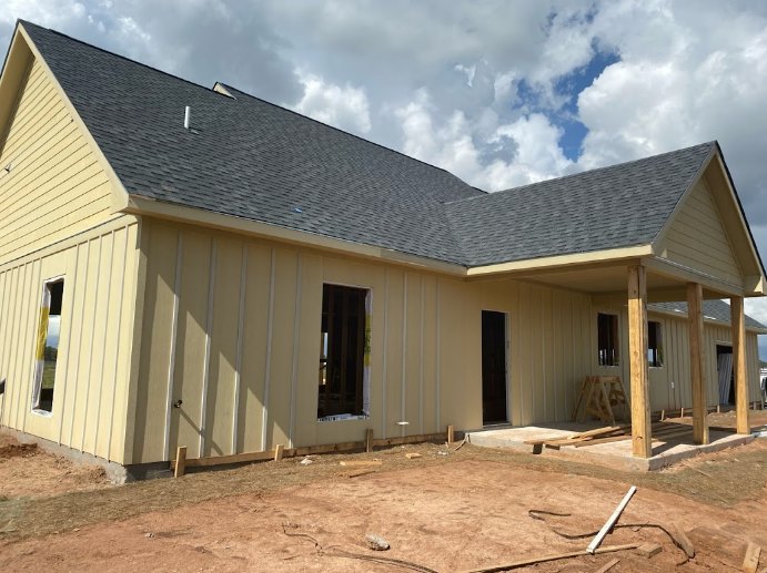 Partially built house with exposed framing, unfinished siding, and dirt yard under cloudy sky