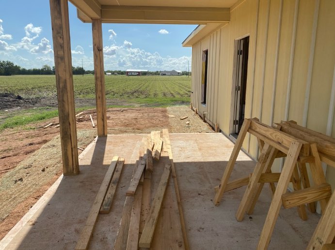 Wood-framed custom home under construction with exposed lumber, concrete floor, sawhorses on the porch, and open field in the background