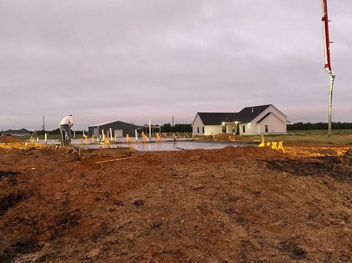 Modern house with illuminated windows, surrounded by dirt and grass, man standing near soil pile, cloudy sky overhead, small body of water in background