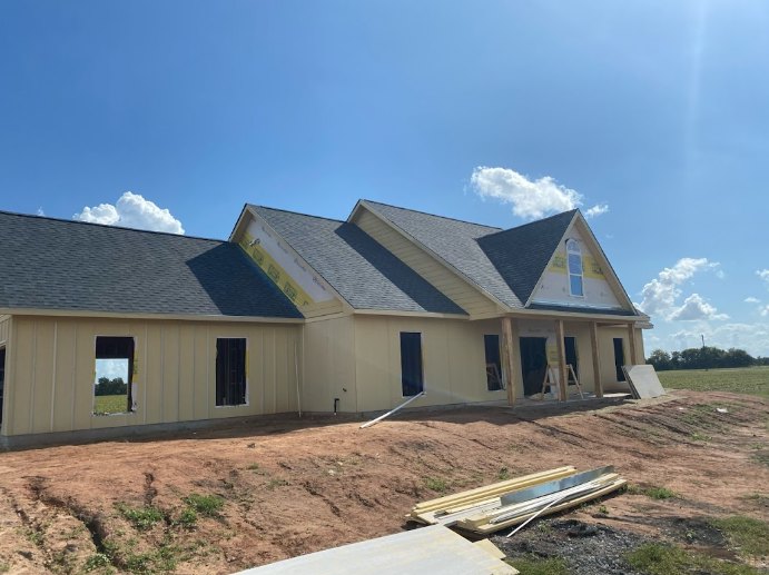 Framed house under construction with exposed roof, dirt hill in foreground, scattered wood and metal materials, blue sky with clouds overhead