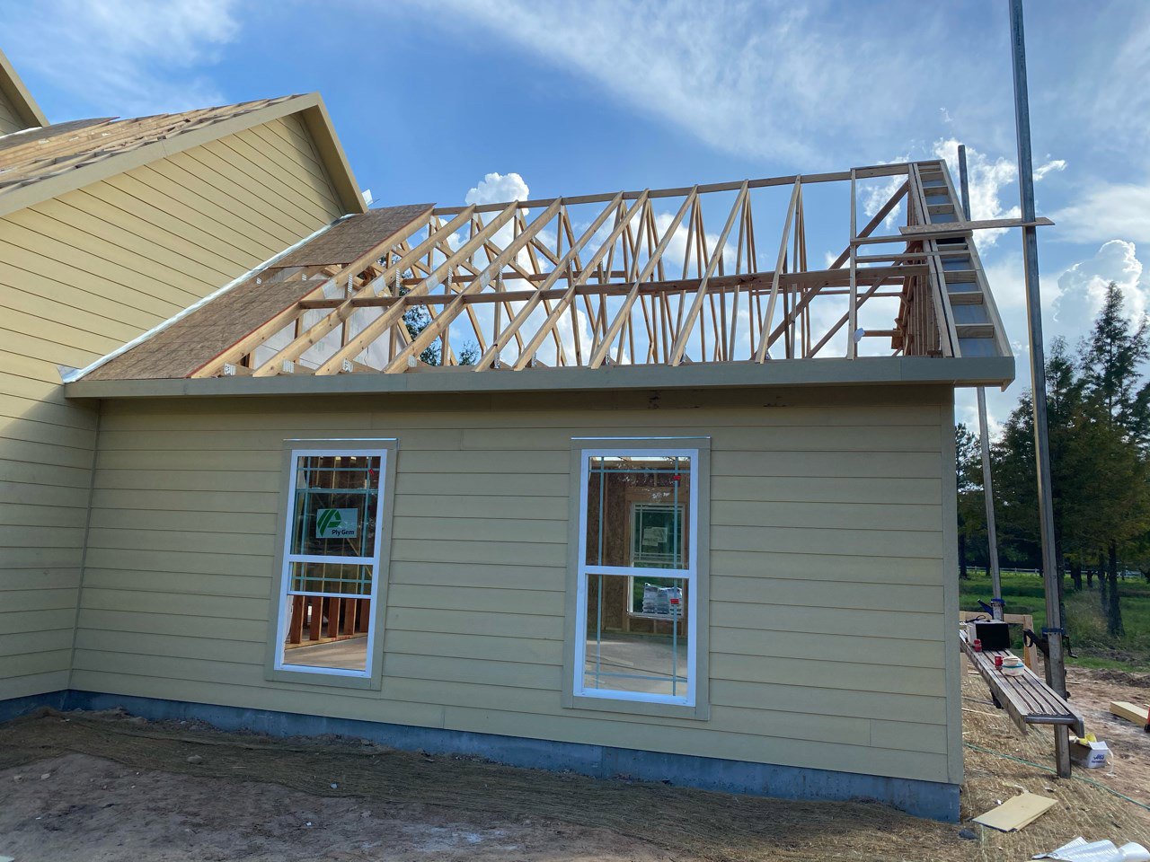 Wood-framed house under construction with white-framed window displaying a sign featuring a green triangle and black text, ladder leaning against unfinished exterior, cloudy sky in