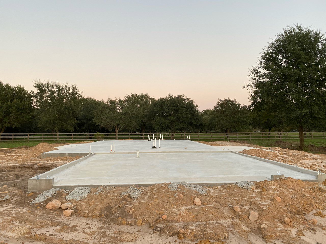 Concrete slab foundation set in a dirt clearing, surrounded by leafy trees under a blue sky