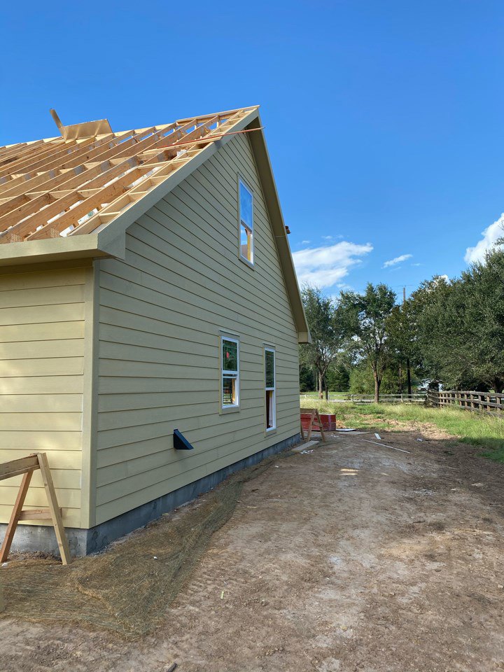 Framed house under construction with exposed wooden beams, white siding, roof partially covered, wooden ladder leaning against wall, surrounded by trees under clear blue sky