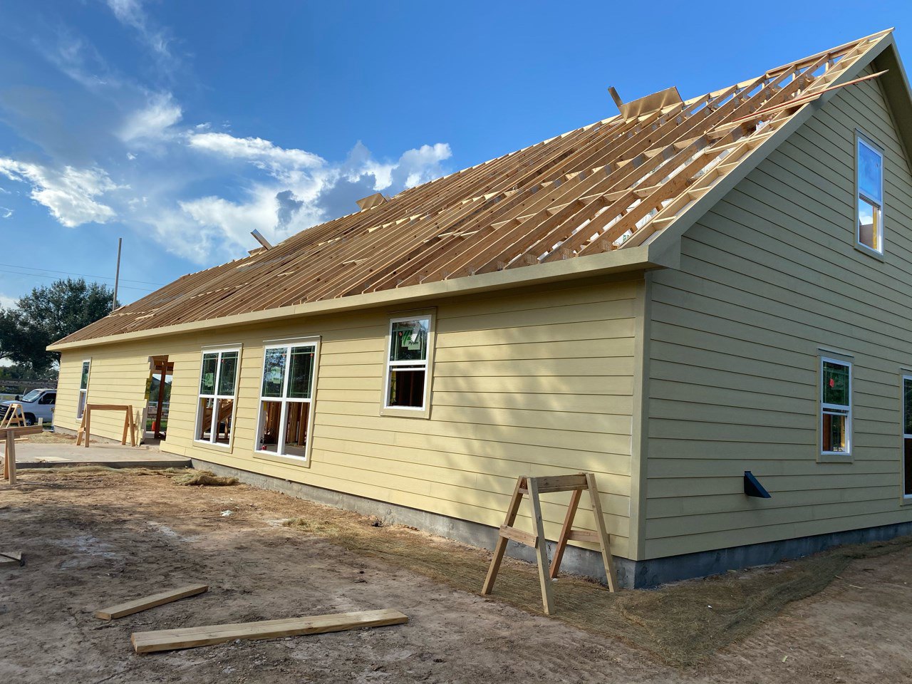 Yellow siding house under construction with a wooden ladder leaning against the wall, white-framed window displaying a sign, white truck parked nearby, cloudy sky overhead.