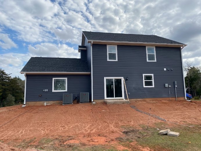 Partially built house with exposed framing, white window frames, ladder leaning against exterior, dirt field in foreground, blue sky overhead