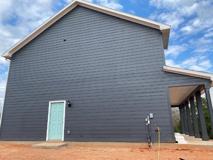 Two-story home with white siding, blue front door with white trim, covered porch supported by white pillars, gray shingle roof, dirt yard, and blue sky with scattered clouds