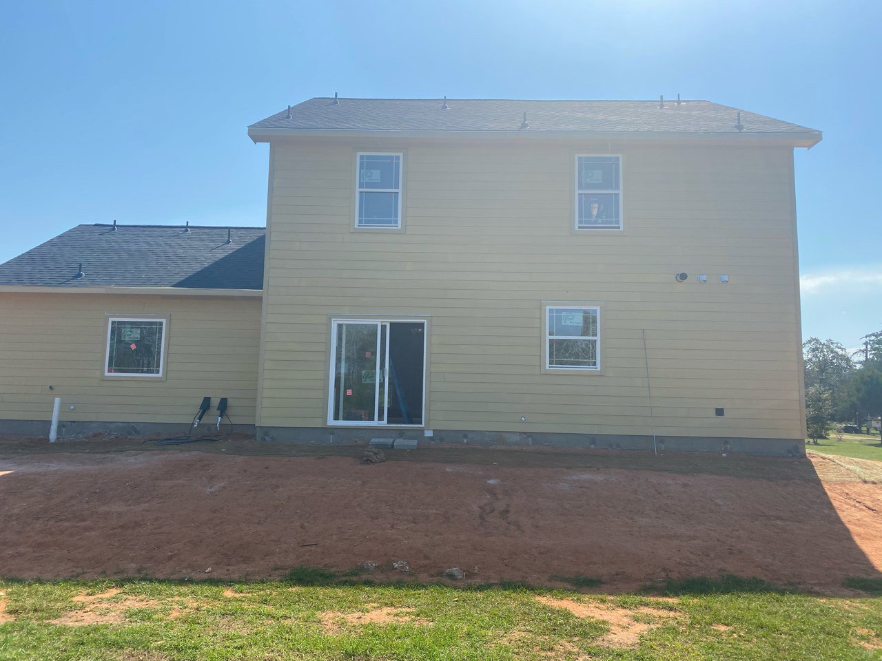 Modern house under construction featuring gray siding, multiple white-framed windows with stickers, sliding glass door, and unfinished dirt yard in front