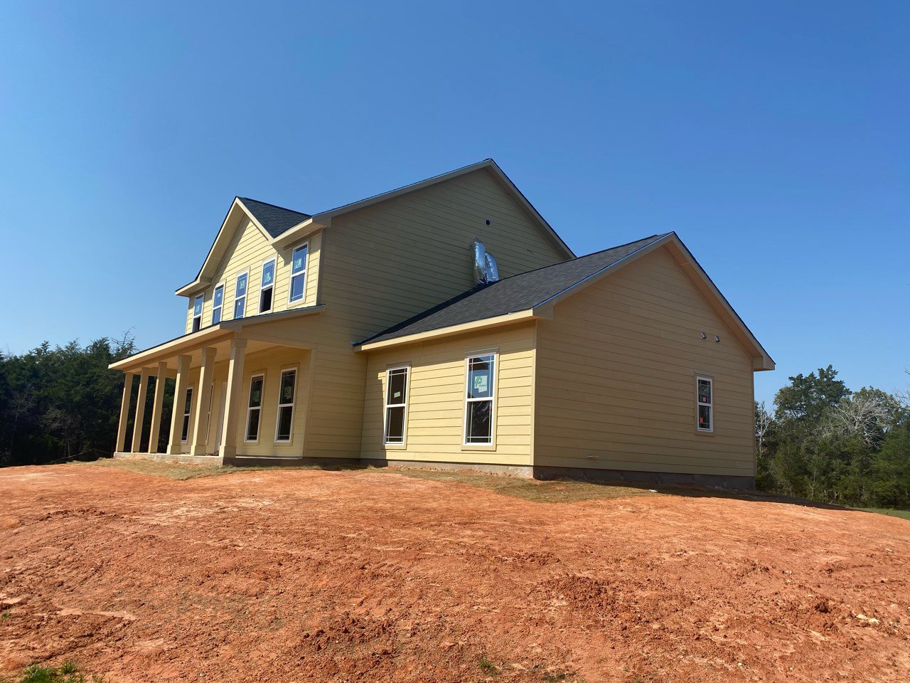Partially built house with exposed framing, dirt yard, leafless tree, roof vent, window displaying construction sign, and clear blue sky