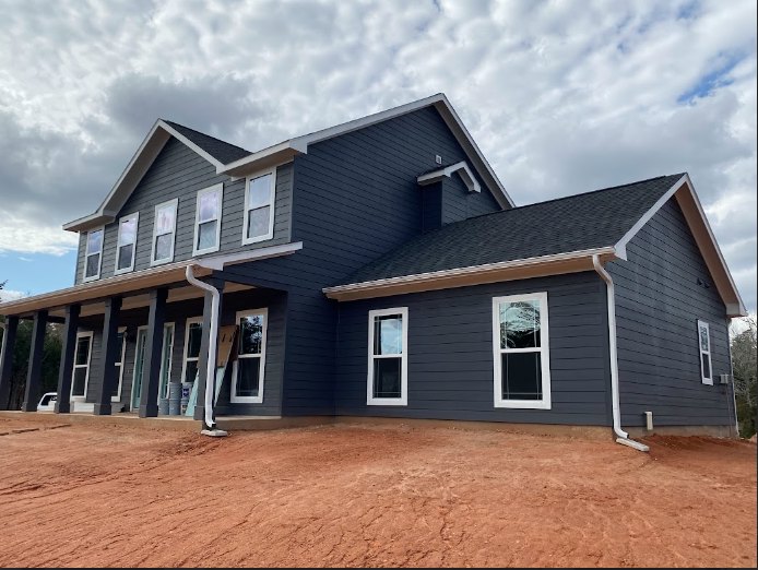 Partially built home with exposed framing, unfinished siding, covered porch, and multiple windows beneath an overcast sky