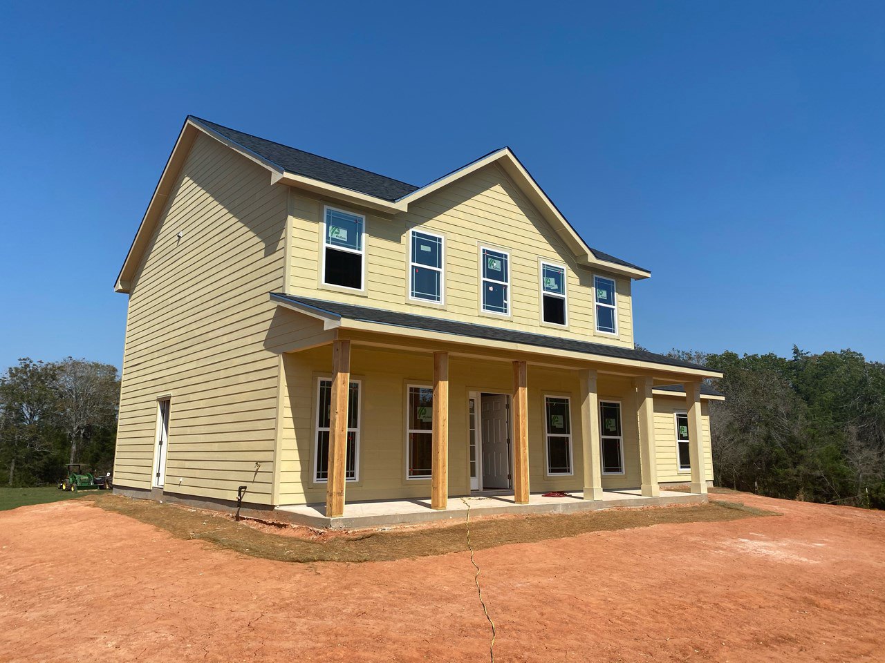 Partially built house with exposed framing, white window frames, and unfinished siding, surrounded by dirt and construction equipment under a clear blue sky