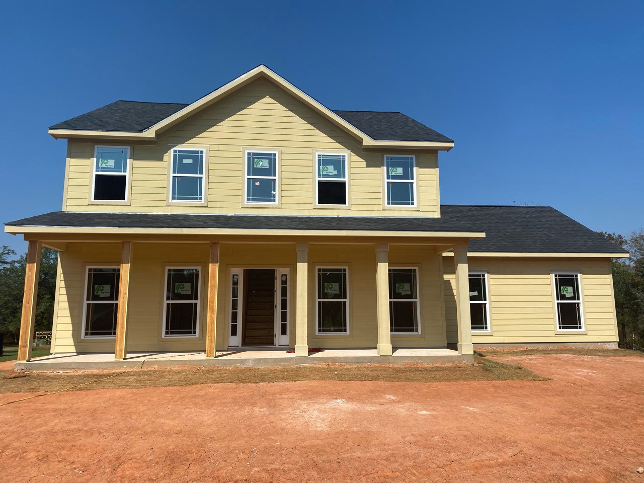 Two-story house under construction with exposed wooden porch, dirt driveway, white siding, roof shingles, and a window displaying a sign against a clear blue sky