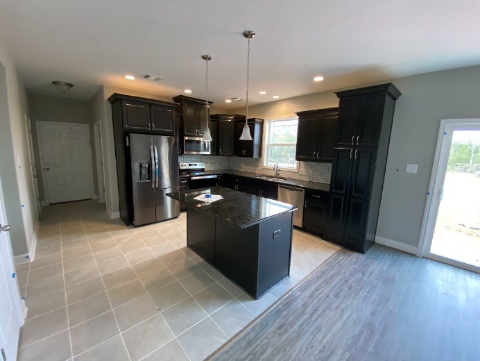 Modern kitchen featuring matte black cabinets, matching black island with waterfall countertop, stainless steel refrigerator, white tile backsplash, light wood flooring, and
