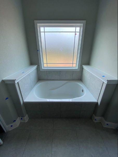 White freestanding bathtub beside frosted glass window, surrounded by white tile walls and square tile flooring with white grout in modern bathroom.