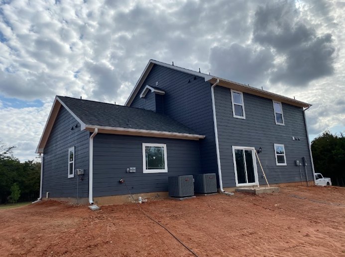 Wood-framed house under construction with exposed beams and unfinished walls, surrounded by trees under a cloudy sky