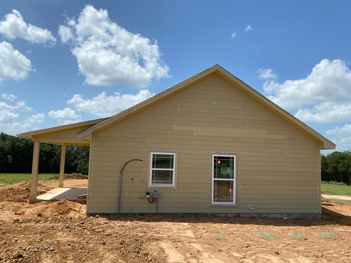 Partially built house with exposed wooden framing, white window frame displaying a sign, dirt lot in foreground, long pipe and electrical wires running along exterior, under clear