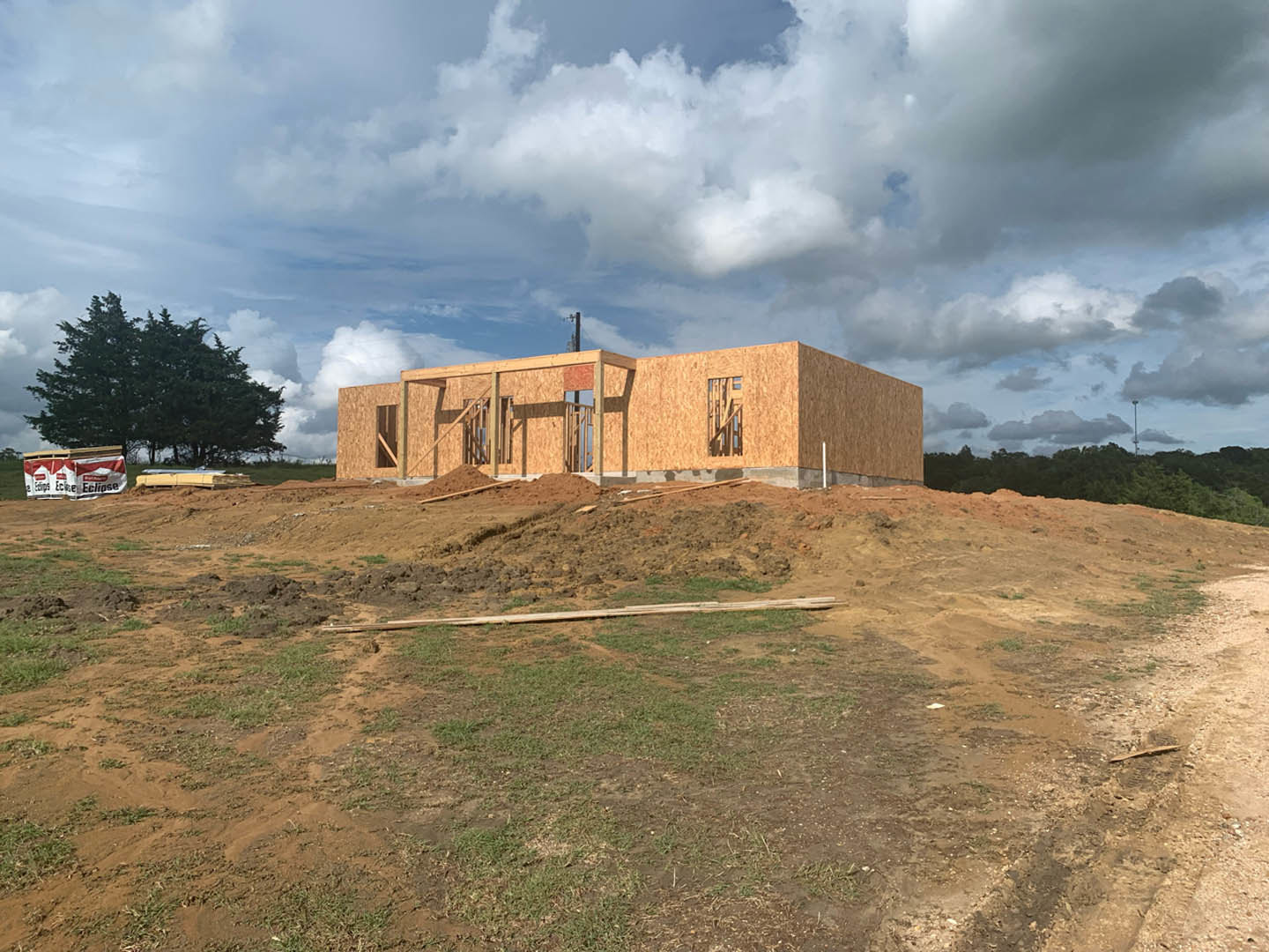 Wood-framed house under construction on a grassy hill, surrounded by trees, dirt piles, and scattered lumber, with a white and red awning displaying black text and clouds overhead.