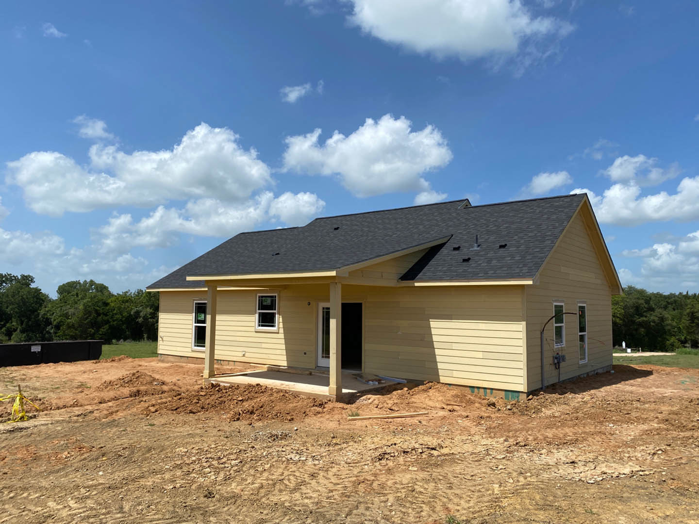 Wood-framed house under construction with exposed plywood walls, dirt lot in foreground, and clear blue sky overhead