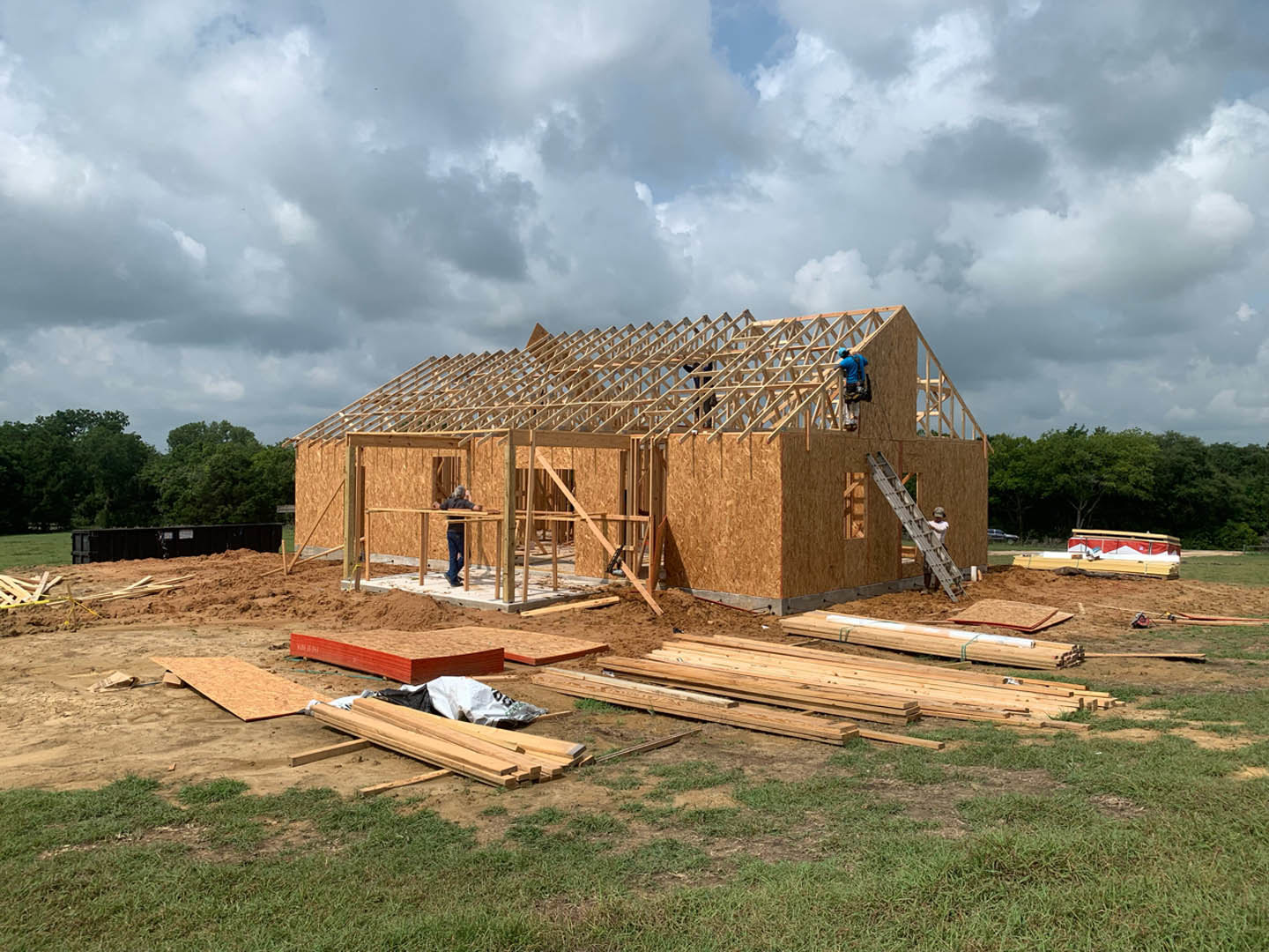 Wood-framed house under construction with exposed beams, people working on site, ladder leaning against structure, black container with white signage, grassy lot and trees in